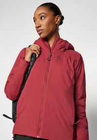 Veste isolée rouge avec capuche, fermeture éclair et poignets élastiques. Présente un logo sur la poitrine et une texture lisse et légère.
