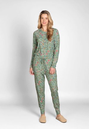 Woman standing wearing green floral long-sleeve top and matching pants with beige slippers on a plain light background.