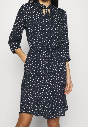 Marineblaues knielanges Kleid mit weißen unregelmäßigen Polka-Dots, dreiviertel Ärmeln, Knopfleiste vorne, Taillengürtel und gebundenem Halsausschnitt.