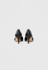 Zapatos de tacón alto de cuero negro con punta puntiaguda y diseño recortado. Presentan un tacón de aguja delgado y un logotipo en tono plateado en la parte trasera.