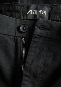 Schwarze Hose aus glattem Stoff mit Knopfverschluss, Reißverschluss und einem Etikett am Bund. Sichtbare Nähte und Taschendetails.