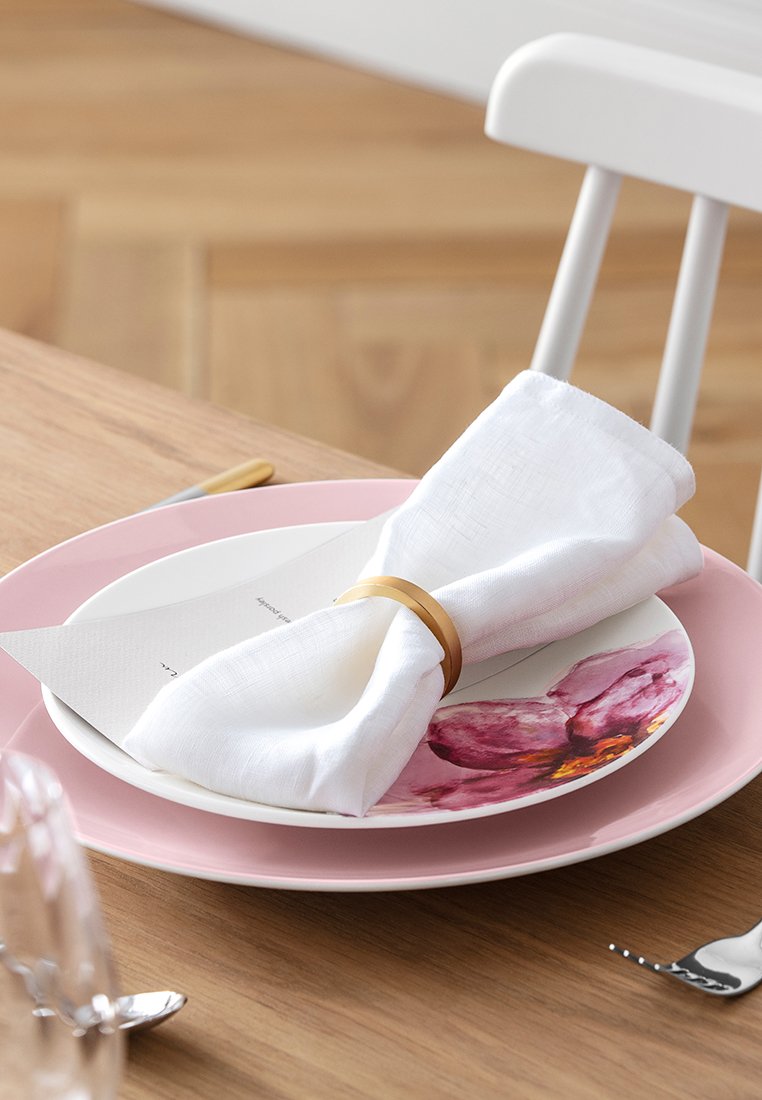 Biała lniana serwetka owinięta złotym pierścieniem na różowym talerzu w kwiatowe wzory, położona na drewnianym stole z sztućcami.