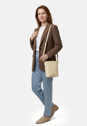 Frau steht und trägt ein braunes Blazer, ein weißes Hemd, hellblaue Hose, beige Mokassins und trägt eine beige Leder-Umhängetasche.