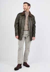 Dunkelgrüne Lederjacke mit einem braunen Fellkragen, zwei Fronttaschen, getragen über einem beigen Rollkragenpullover, kombiniert mit hellgrauen Jeans und braunen Stiefeln.