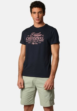T-shirt en coton bleu marine avec un texte graphique rouge, associé à un short cargo vert clair. La tenue est décontractée et convient aux temps chauds.