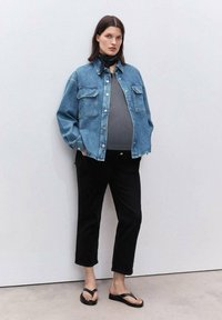 Giacca di jeans azzurra con vestibilità comoda, top grigio, pantaloni neri a vita alta e infradito neri. Modello in piedi contro uno sfondo neutro.