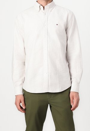 Mężczyzna w białej koszuli z długim rękawem zapinanej na guziki z cienkimi pionowymi paskami i oliwkowozielone spodnie na jednolitym tle.