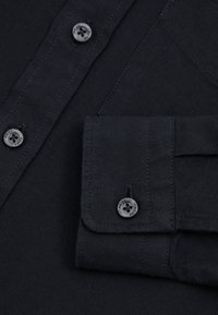 Close-up af en sort skjortemanchet og knapstolpe med grå knapper mærket "REISS", der viser stofstruktur og syningsdetaljer.