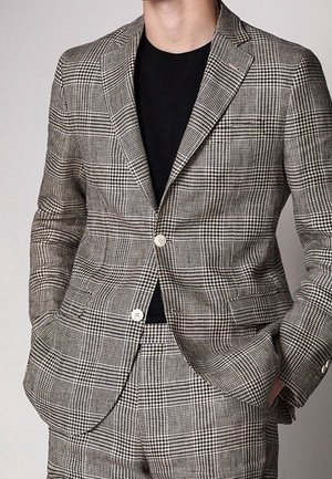 Homme portant un costume à motif pied-de-poule gris et noir avec une chemise noire, les mains dans les poches, tête non visible.