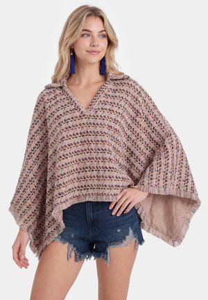 Donna bionda che indossa un poncho multicolore con fantasia, con frange e orecchini con nappine blu, abbinato a pantaloncini di jeans strappati, posando con una mano sul fianco.