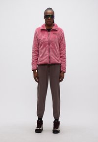 Άτομο που στέκεται φορώντας ροζ φλις ζακέτα με φερμουάρ, καφέ παντελόνι jogging, πεζοπορικά μποτάκια και αντανακλαστικά γυαλιά ηλίου μπροστά σε απλό φόντο.