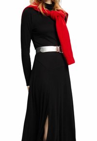 Schwarzes Kleid mit langen Ärmeln und einem Seitenschlitz, kombiniert mit einem roten Strickschal und einem silbernen metallischen Gürtel, der eine glatte Textur aufweist.