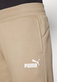 Šviesiai rudos spalvos treningo kelnės, pagamintos iš minkštos medžiagos, su rombuotu juosmeniu ir šoninėmis kišenėmis. Priekyje esantis baltas Puma logotipas.
