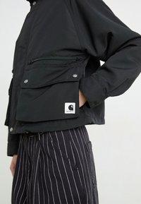 Čierna nylónová bunda so zipsom a tlačným gombíkovým detailom, s logom na dolnej ľavej strane. V páre so pruhovanými nohavicami.