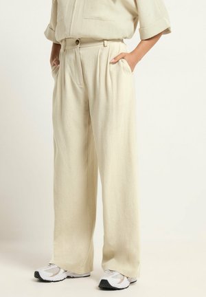 Beżowe szerokie spodnie z wysokim stanem, z zakładkami, zapinane na guzik i zamek, Noszone z białymi trampkami i rękami w kieszeniach.