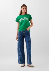 T-shirt verde in cotone con testo bianco "SCALPERS", abbinato a jeans in denim blu a gamba larga con un dettaglio di cucitura distintivo. Sneakers bianche completano l'outfit.