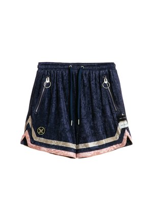 Joven mujer con cabello rubio está de pie vistiendo una camisa azul marino de botones de gran tamaño y pantalones cortos a juego con rayas multicolor, calcetines blancos y zapatillas beige.