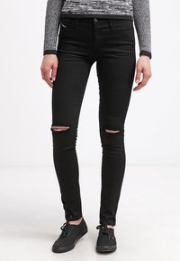 Jeans negros ajustados con rodillas rasgadas, que presentan una textura suave y un corte slim. Combinados con zapatillas negras para crear contraste.
