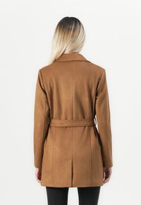 Femme aux longs cheveux blonds portant un manteau marron ceinturé, tournée vers une arrière-plan blanc uni.