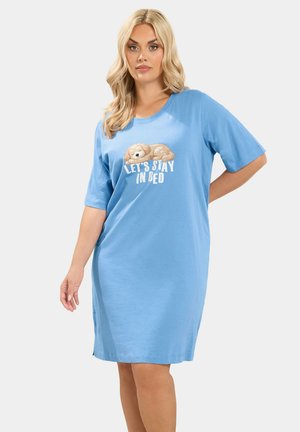 Kvinde med langt blond hår iført lyseblå natkjole med illustration af sovende hund og tekst "LET'S STAY IN BED" på fronten.