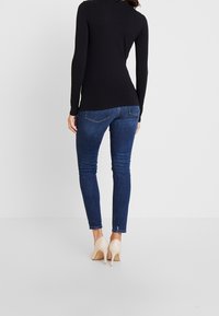 Schwarz langärmliges Oberteil, kombiniert mit dunkelblauen engen Jeans und nudefarbenen High Heels. Das Material wirkt glatt mit einer figurbetonten Silhouette.
