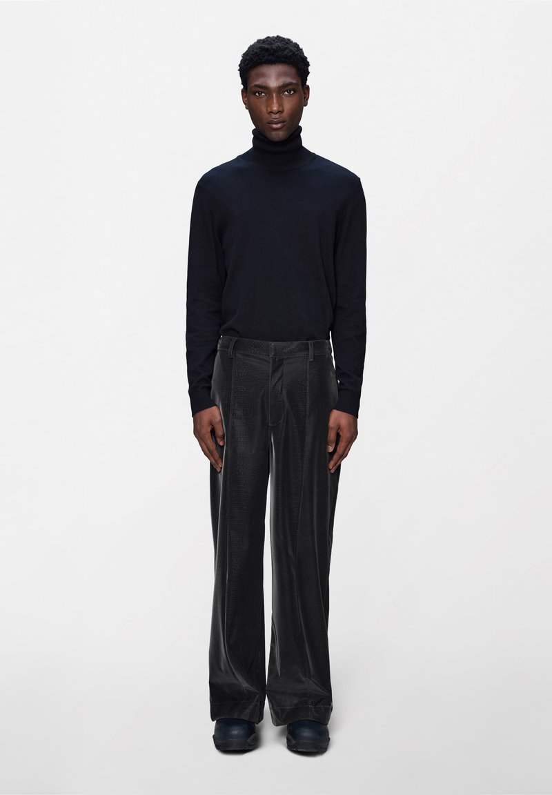 Mees seismas ühevärvilise tausta ees, seljas must kõrge kaelusega särk, laiad tekstuuriga mustad püksid ja mustad kingad, vaadates otse ette, käed kõrval.