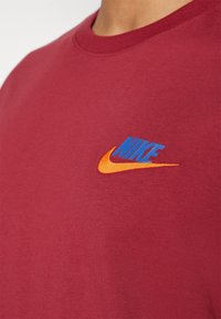Gros plan d'un t-shirt rouge avec un logo Nike bleu et un swoosh orange sur la poitrine, porté par une personne avec un cou visible.