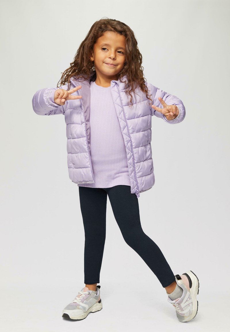 Jasnofioletowa kurtka puchowa, żeberkowy lawendowy top, czarne legginsy i sportowe sneakersy z pastelowymi akcentami. Modelka przybiera pozę z znakiem pokoju.