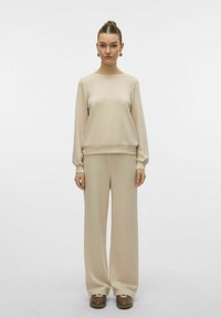 Felpa beige e pantaloni larghi realizzati in tessuto morbido. Presenta un collo rotondo e polsini a coste. Il modello sta in piedi con le mani ai lati.