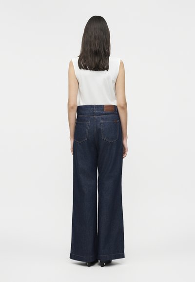 Γυναίκα με σκούρα μαλλιά μέχρι τους ώμους, ντυμένη με λευκό αμάνικο τοπ και φαρδιά σκούρα μπλε τζιν, στέκεται με την πλάτη προς τα εμπρός σε λευκό φόντο.