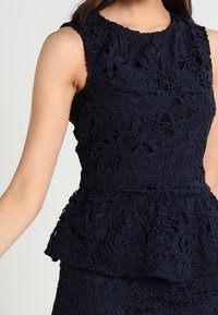 Top peplum senza maniche di colore blu navy, realizzato in pizzo testurizzato con motivi floreali, scollo rotondo e silhouette aderente.