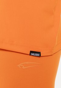 Oranje sportbroekje gemaakt van gladde stof met een zwart "SMILODOX" label en een lichtoranje logo aan de zijkant.