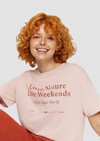 Roze katoenen t-shirt met een relaxte pasvorm, voorzien van rode tekst: "Hou van de natuur, leef in het weekend" en "Ken je waarde." Korte mouwen en een zachte textuur.