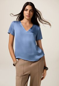 Donna con i capelli lunghi e scuri, che indossa una blusa azzurro chiaro a scollatura a V e pantaloni beige, con le mani nelle tasche, in posa su uno sfondo neutro.