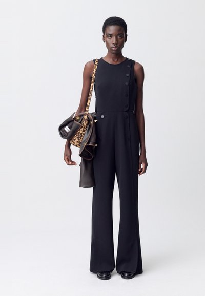 Femme portant une combinaison noire sans manches avec des détails de boutons, tenant une veste en cuir et un sac à imprimé léopard avec une bandoulière sur l'épaule.