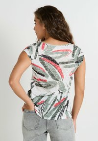 Top de manga corta en tela ligera con un patrón de hojas verdes, rojas y negras sobre un fondo blanco. Escote redondeado con un sutil brillo.