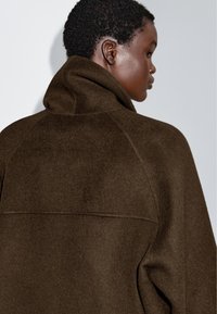 Manteau en laine marron avec un col haut et des manches raglan, présentant une finition texturée et un design minimaliste. La vue arrière révèle des détails de couture.