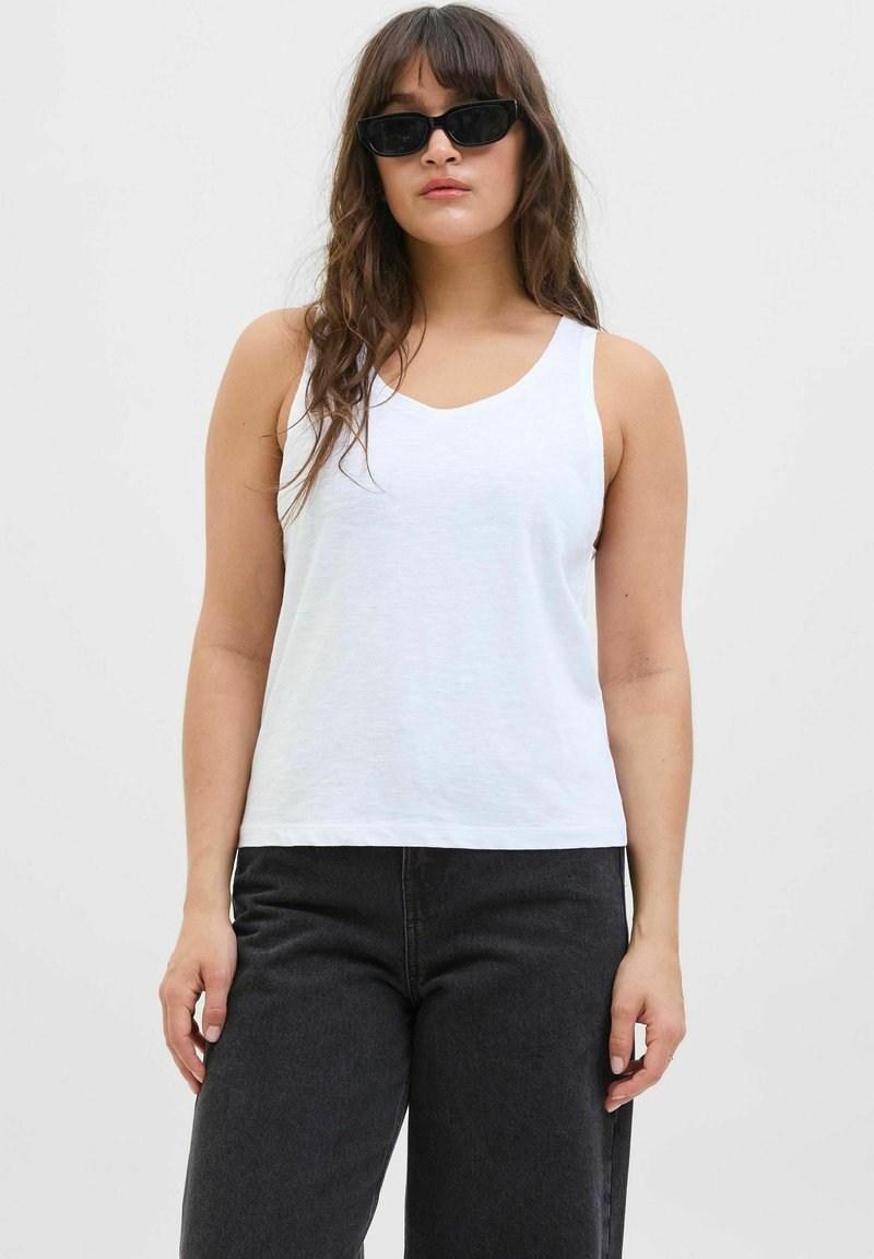 Top sin mangas blanco con un diseño de cuello en V, hecho de una tela suave. Combinado con pantalones oscuros de cintura alta en gris.