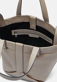 Beżowa skórzana torba tote o wyraźnym kształcie. Posiada wnętrze z kieszenią na zamek błyskawiczny i czarne materiałowe wykończenie, uzupełnione długimi, mocnymi uchwytami.