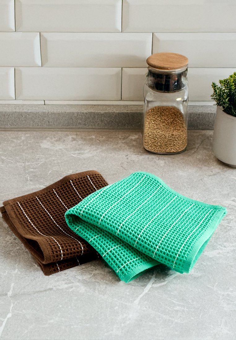 Na zdjęciu widać dwa ściereczki kuchenne: jedna brązowa z teksturowanym wzorem i białymi paskami, a druga jasnozielona z podobnym wzorem, umieszczone na szarym blacie.