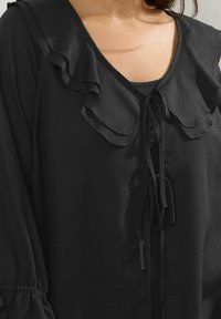 Svart blus med volangkrage, dragsko-stängning och texturerad tyg. Långa ärmar med samlad detalj vid manschetterna.