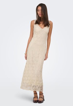 Mujer con un vestido largo de encaje beige con tirantes finos y zapatos Mary Jane marrones, de pie frente a un fondo liso y claro.