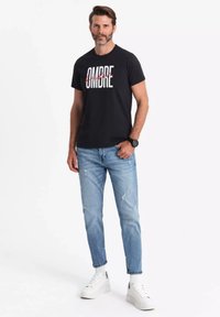 Camiseta negra de cuello redondo con gráfico "OMBRE" en blanco y rojo. Combinada con jeans de mezclilla destrozados de color azul claro y zapatillas blancas.