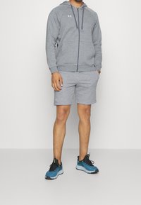 Sudadera gris con cremallera, con un bolsillo tipo canguro y pantalones cortos a juego, combinados con zapatillas atléticas azules que tienen detalles y cordones verdes.