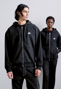 Čierne mikiny s kapucňou na zips a prednými vreckami. Na prednej strane sa nachádza malá logo. Jeden model má na sebe ošúchané čierne džínsy s bielym prešívaním.