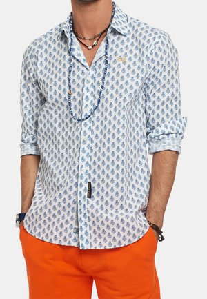 Mand iført en hvid skjorte med blå blomsterprint, orange bukser, perlehalskæder, et ur og armbånd, med hænderne i lommerne.