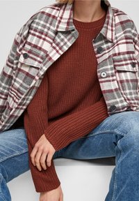 Geruit overhemd in grijs, wit en bordeaux, gedragen over een gebreide trui met een geribbelde roestkleurige textuur. Lichtblauwe spijkerbroek met geweven details.