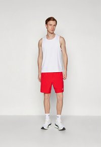 Witte mouwloze tanktop en rode sportshorts. De outfit heeft een klassiek ontwerp met een logo op de shorts. Draagt witte sneakers.