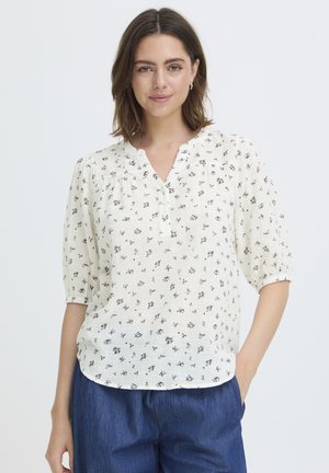 Kvinde med skulderlangt brunt hår iført en hvid blomstret bluse med knapper og cowboybukser, stående med den ene hånd i lommen.