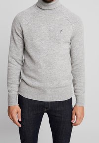 Homme portant un pull à col roulé gris clair avec un petit logo d'oiseau sur la poitrine et un jean bleu foncé, se tenant devant un fond uni.